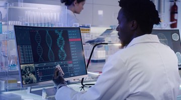 Photo: Researcher at a computer looking at a sample in hand as well as computer screen, which shows DNA.